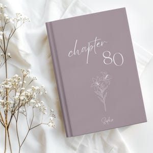 May include: A dusty mauve hardcover book featuring the white text "chapter 80" and a delicate line art illustration of two lilies. The name "Sophie" is printed in a cursive script at the bottom center of the front cover of the book.