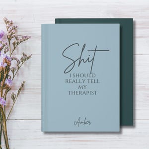 May include: A light blue notebook with dark gray text that reads "Shit I SHOULD REALLY TELL MY THERAPIST" and the name "Amber" at the bottom. The notebook is positioned in front of a dark green cover, with a sprig of purple flowers to the left.