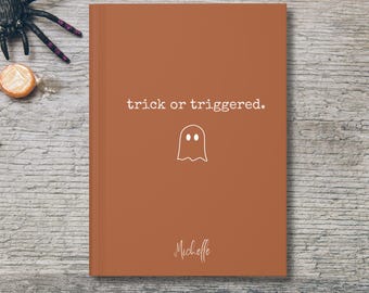 May include: A burnt orange journal with the text "trick or triggered" and a ghost illustration. The name "Michelle" is written in cursive at the bottom. The journal is on a wooden surface with Halloween decorations.