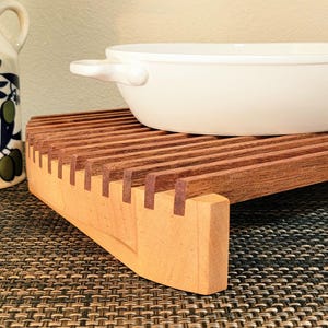 May include: A rectangular wooden trivet featuring a slatted design with dark brown horizontal slats and light tan wooden end supports. A white oval ceramic baking dish with two small side handles rests on top of the wooden trivet structure.