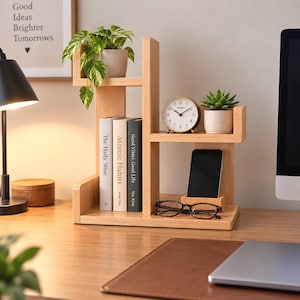 May include: A light-colored wooden desktop organizer with multiple tiers. The unit holds three books, a small potted pothos plant, a round analog clock, a smartphone in a dedicated slot, and a pair of tortoiseshell-rimmed eyeglasses on the base.