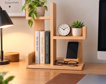 May include: A light-colored wooden desktop organizer with multiple tiers. The unit holds three books, a small potted pothos plant, a round analog clock, a smartphone in a dedicated slot, and a pair of tortoiseshell-rimmed eyeglasses on the base.