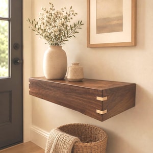 May include: A rectangular floating wall shelf made of dark brown wood with visible light-colored dovetail joinery on the corners. A large beige ceramic vase with white flowers and a smaller matching beige ceramic jar sit on the shelf surface.