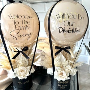 May include: Two hot air balloon arrangements with white flowers and pampas grass in black boxes. The balloons are clear with gold confetti and black lettering. One balloon says "Welcome to the Family Sunny" and the other says "Will You Be Our Dhulabhai". The boxes have dates on them: 20.10.24 and 27th October 2024.