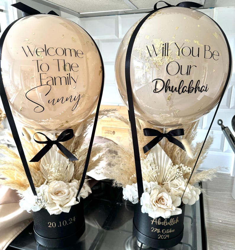 May include: Two hot air balloon arrangements with white flowers and pampas grass in black boxes. The balloons are clear with gold confetti and black lettering. One balloon says "Welcome to the Family Sunny" and the other says "Will You Be Our Dhulabhai". The boxes have dates on them: 20.10.24 and 27th October 2024.