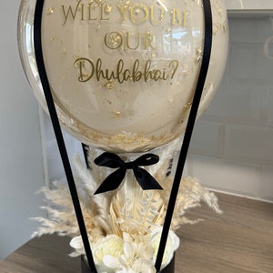 May include: A clear balloon hot air balloon with gold confetti and a black ribbon. The balloon has a gold inscription that reads "Will you be our Dhulabhai?" The balloon is attached to a black box with white flowers and dried pampas grass. The box has a gold inscription that reads "Love from all your Hola & Hel's"