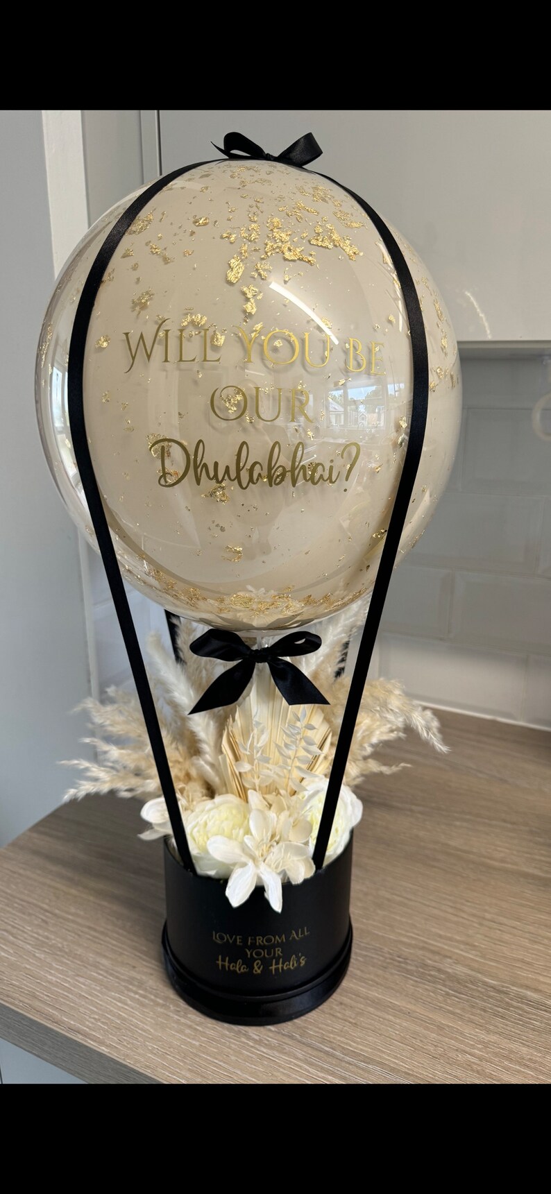 May include: A clear balloon hot air balloon with gold confetti and a black ribbon. The balloon has a gold inscription that reads "Will you be our Dhulabhai?" The balloon is attached to a black box with white flowers and dried pampas grass. The box has a gold inscription that reads "Love from all your Hola & Hel's"