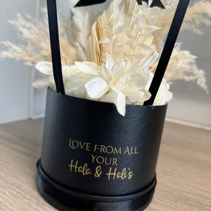 May include: A black gift box with a white ribbon and dried flowers. The box has a gold inscription that reads "Love from all your Hala & Hali's".