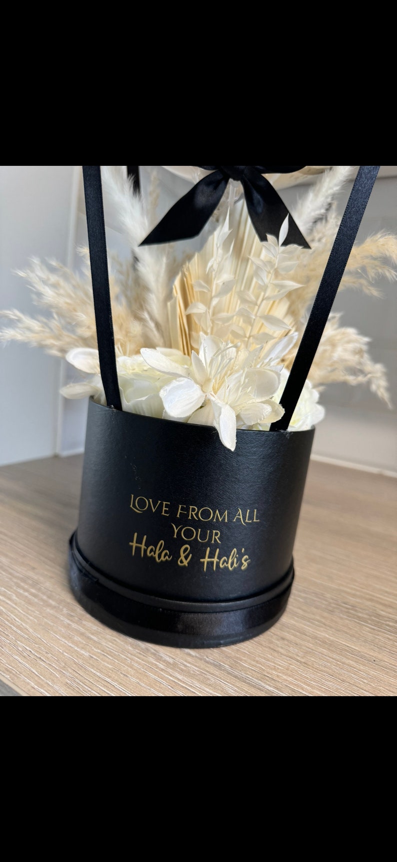 May include: A black gift box with a white ribbon and dried flowers. The box has a gold inscription that reads "Love from all your Hala & Hali's".