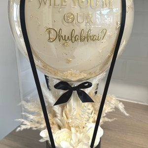 May include: A clear glass balloon hot air balloon with gold flecks and a black ribbon tied around the bottom. The balloon has a gold inscription that reads "Will you be our Dhulabhai?" The balloon is attached to a black box with white dried flowers and a gold inscription that reads "Love from all your Hola & Hel's".