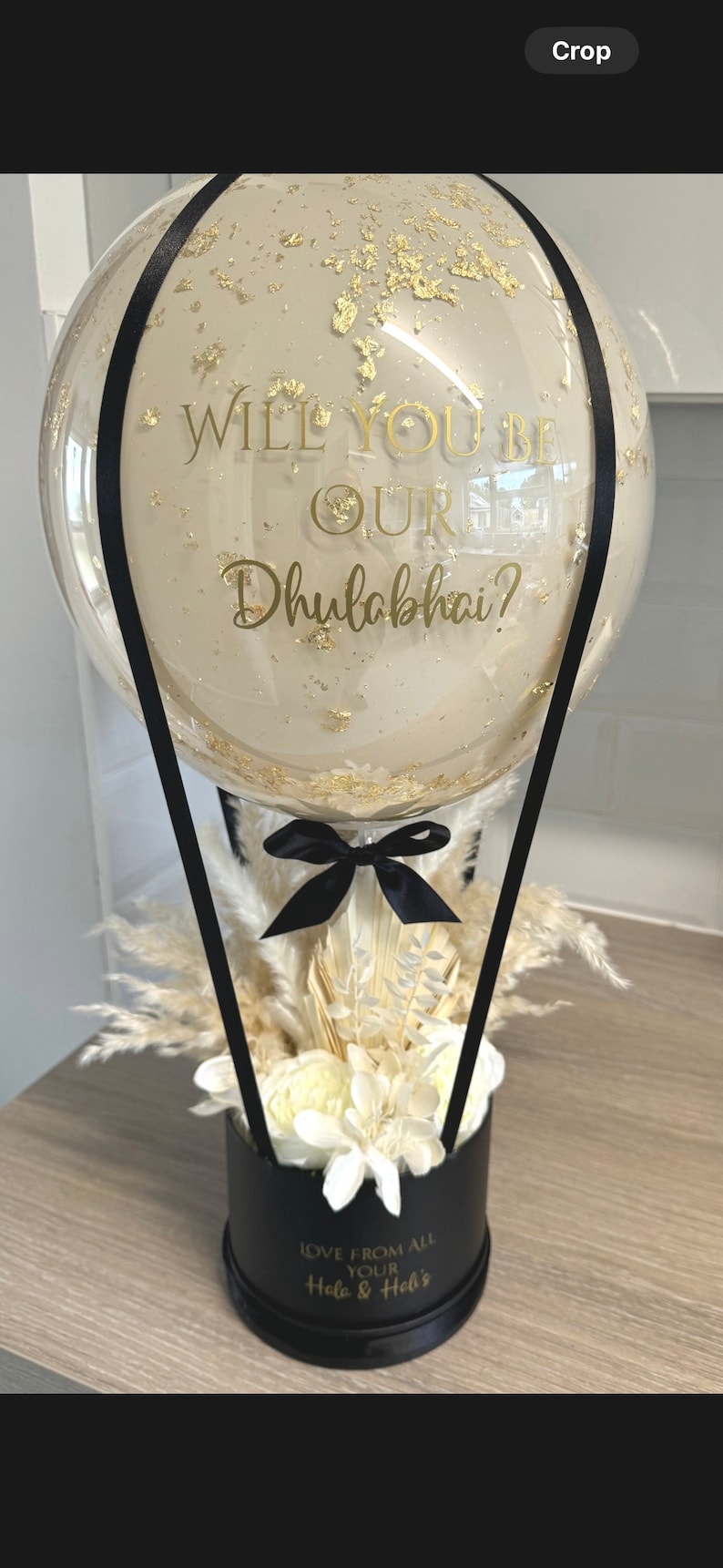 May include: A clear glass balloon hot air balloon with gold flecks and a black ribbon tied around the bottom. The balloon has a gold inscription that reads "Will you be our Dhulabhai?" The balloon is attached to a black box with white dried flowers and a gold inscription that reads "Love from all your Hola & Hel's".
