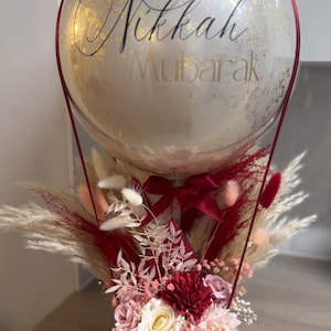 May include: A red box filled with dried flowers and greenery, topped with a clear balloon with gold glitter and the text "Nikkah Mubarak".