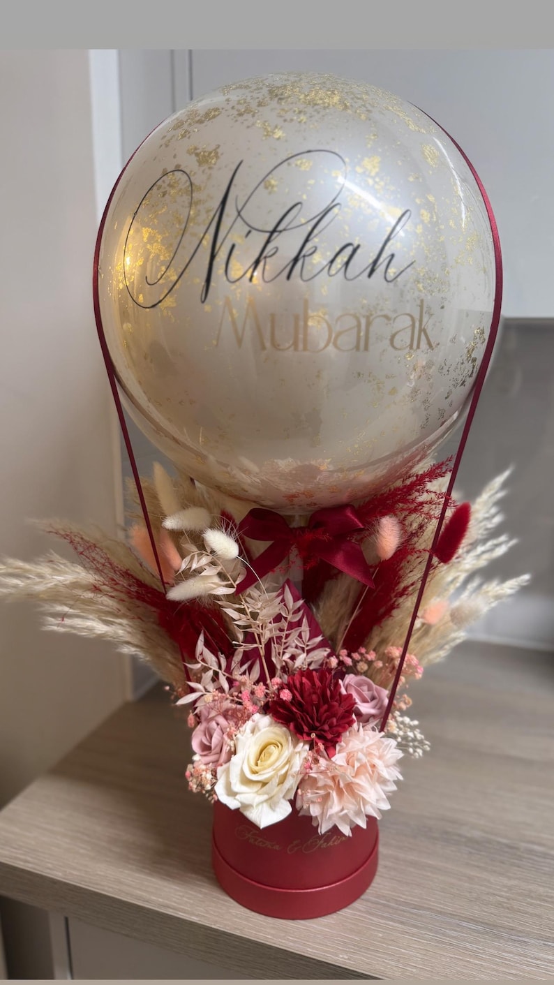 May include: A red box filled with dried flowers and greenery, topped with a clear balloon with gold glitter and the text "Nikkah Mubarak".