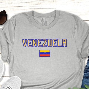 May include: A gray t-shirt with the word 'VENEZUELA' in blue and yellow letters. A small Venezuelan flag is below the word. The t-shirt is laid out on a wooden surface with a pair of white sneakers, blue jeans, and a green plant sprig.