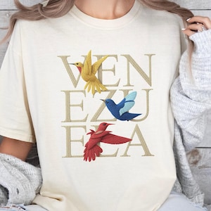 May include: Cream-colored t-shirt featuring the word "VENEZUELA" in gold lettering, with three colorful bird illustrations. The birds are yellow, blue, and red. The shirt is a casual, comfortable style.