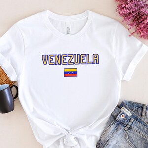 May include: White t-shirt with the word "VENEZUELA" in yellow and blue lettering, and a small Venezuelan flag. The shirt is paired with denim shorts. A black mug and a woven placemat are visible in the background.