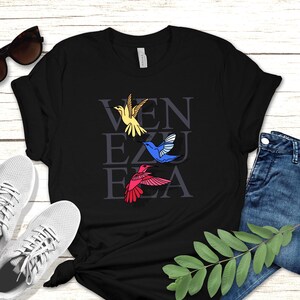 May include: Black t-shirt with the word "VENEZUELA" written in white letters with a yellow, blue, and red hummingbird flying above each letter.