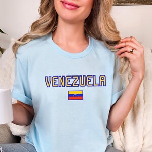 May include: Light blue t-shirt with the word "VENEZUELA" in yellow and blue lettering, and a small Venezuelan flag graphic. The shirt is made of a soft material.