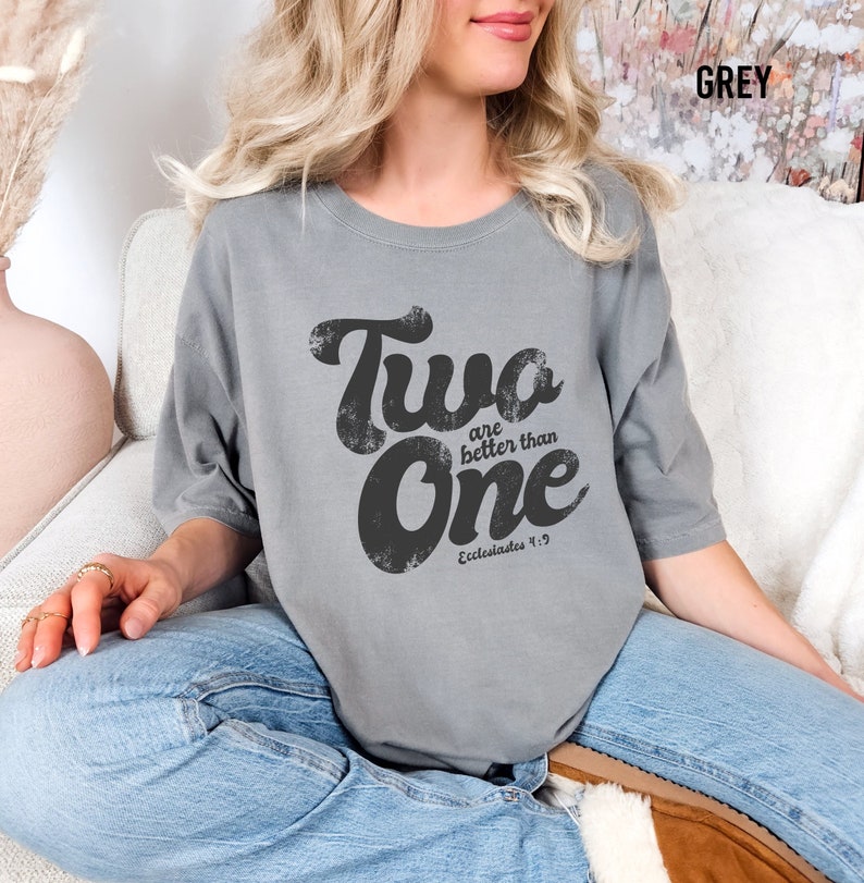May include: A grey short-sleeved t-shirt featuring the black distressed text Two are better than One and the reference Ecclesiastes 4:9 printed on the front. The shirt has a crew neckline and a relaxed fit, paired with blue denim jeans.