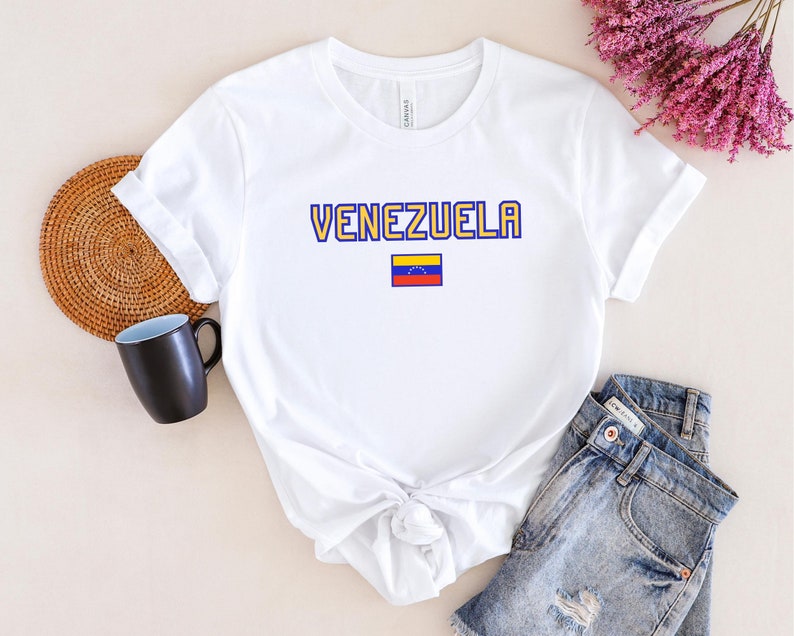 May include: White t-shirt with the word "VENEZUELA" in yellow and blue lettering, and a small Venezuelan flag. The shirt is paired with denim shorts. A black mug and a woven placemat are visible in the background.