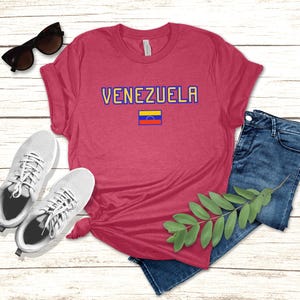 May include: A red t-shirt with the word "VENEZUELA" in blue and yellow lettering, and a small Venezuelan flag. The shirt is paired with blue jeans, white sneakers, and sunglasses.