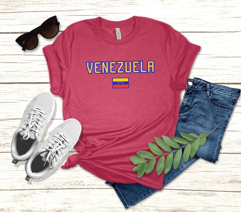 May include: A red t-shirt with the word "VENEZUELA" in blue and yellow lettering, and a small Venezuelan flag. The shirt is paired with blue jeans, white sneakers, and sunglasses.