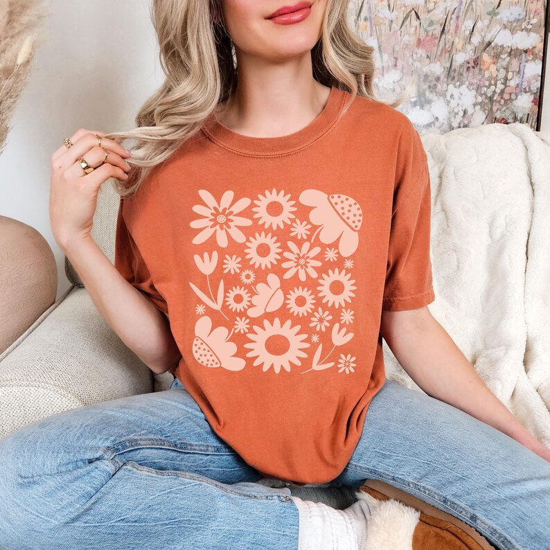 May include: Rust-colored t-shirt featuring a floral design in a lighter shade of peach. The design includes various flower shapes, leaves, and a butterfly. The shirt is a casual, comfortable style.