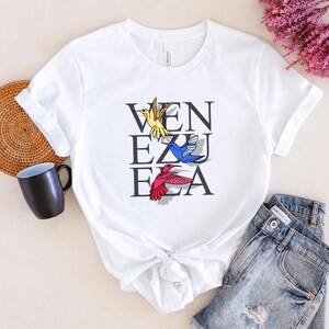 May include: White t-shirt with a graphic of hummingbirds and the word "VENEZUELA" printed on it.