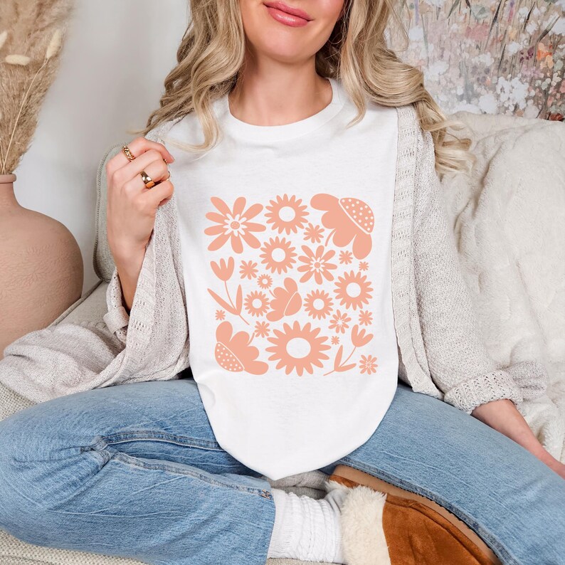 May include: White t-shirt featuring a peach-colored floral design. The design includes various flower shapes and sizes, creating a cohesive pattern. The shirt is part of a casual clothing collection.