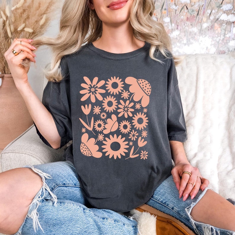 May include: Dark grey t-shirt featuring a peach-colored floral design. The design includes various flower shapes, leaves, and a butterfly. The shirt is a casual style, with a relaxed fit and short sleeves. The model is wearing blue jeans.
