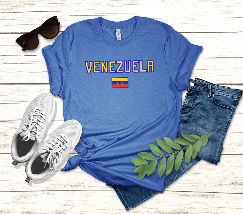 May include: A blue t-shirt with the word 'VENEZUELA' printed in yellow letters. A small Venezuelan flag is printed below the word. The t-shirt is laying on a wooden surface with a pair of white sneakers and a pair of blue jeans.