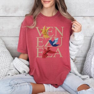 May include: Red t-shirt featuring the word "VENEZUELA" in gold, with illustrations of hummingbirds in yellow, blue, and red. The shirt has a relaxed fit.