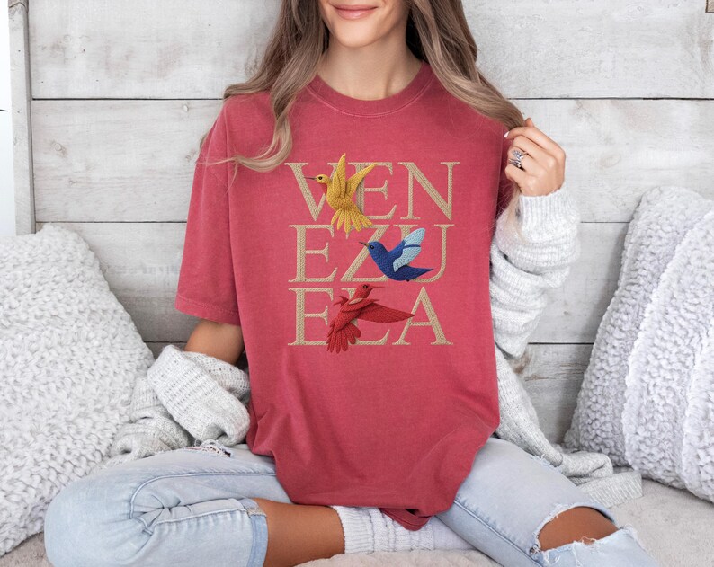 May include: Red t-shirt featuring the word "VENEZUELA" in gold, with illustrations of hummingbirds in yellow, blue, and red. The shirt has a relaxed fit.
