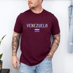 May include: A maroon t-shirt with the word "VENEZUELA" in blue and yellow pixelated letters. Below the text is the Venezuelan flag. The shirt is short-sleeved and worn by a person.