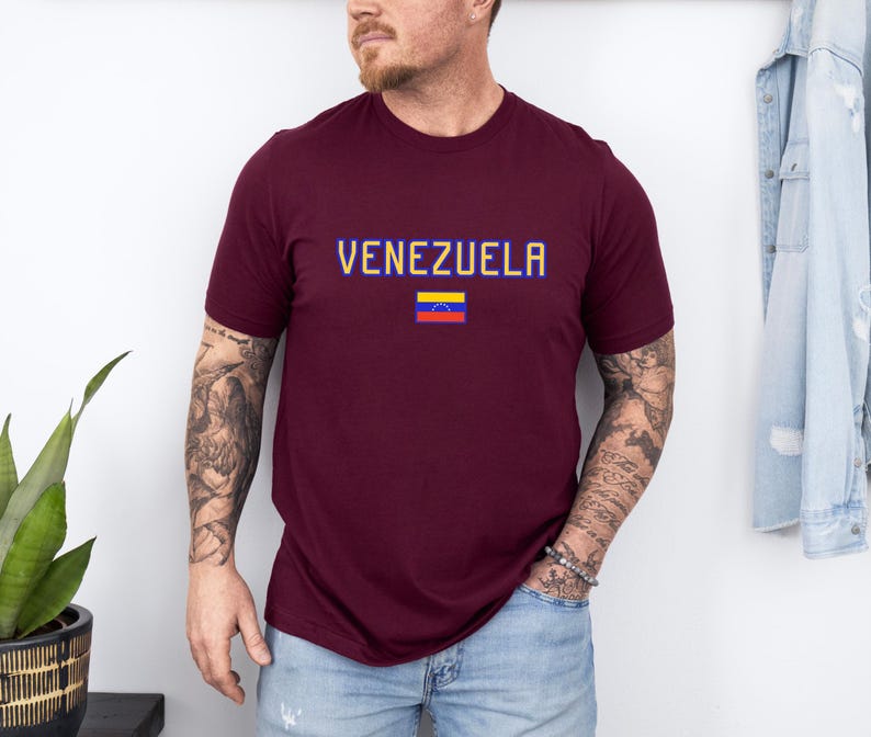 May include: A maroon t-shirt with the word "VENEZUELA" in blue and yellow pixelated letters. Below the text is the Venezuelan flag. The shirt is short-sleeved and worn by a person.