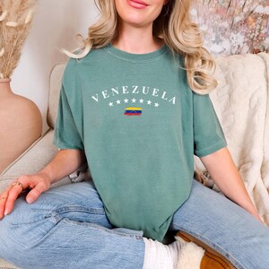Pode incluir: Uma camiseta verde-azulada com a palavra "VENEZUELA" impressa em branco com uma pequena bandeira venezuelana por baixo. A camiseta est&aacute; sendo usada por uma pessoa sentada em uma superf&iacute;cie branca e felpuda.