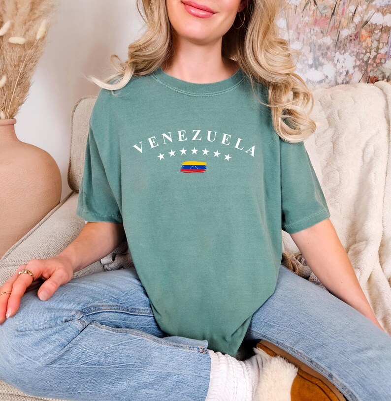 Pode incluir: Uma camiseta verde-azulada com a palavra "VENEZUELA" impressa em branco com uma pequena bandeira venezuelana por baixo. A camiseta est&aacute; sendo usada por uma pessoa sentada em uma superf&iacute;cie branca e felpuda.