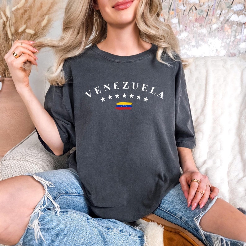May include: A dark gray t-shirt with the word "VENEZUELA" printed in white with a small Venezuelan flag underneath. The shirt has a relaxed fit and is made of a soft, comfortable fabric.