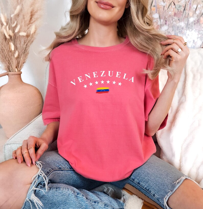 Pode incluir: Uma camiseta rosa com a palavra "VENEZUELA" impressa em branco com uma pequena bandeira venezuelana por baixo. A camiseta est&aacute; sendo usada por uma pessoa sentada.