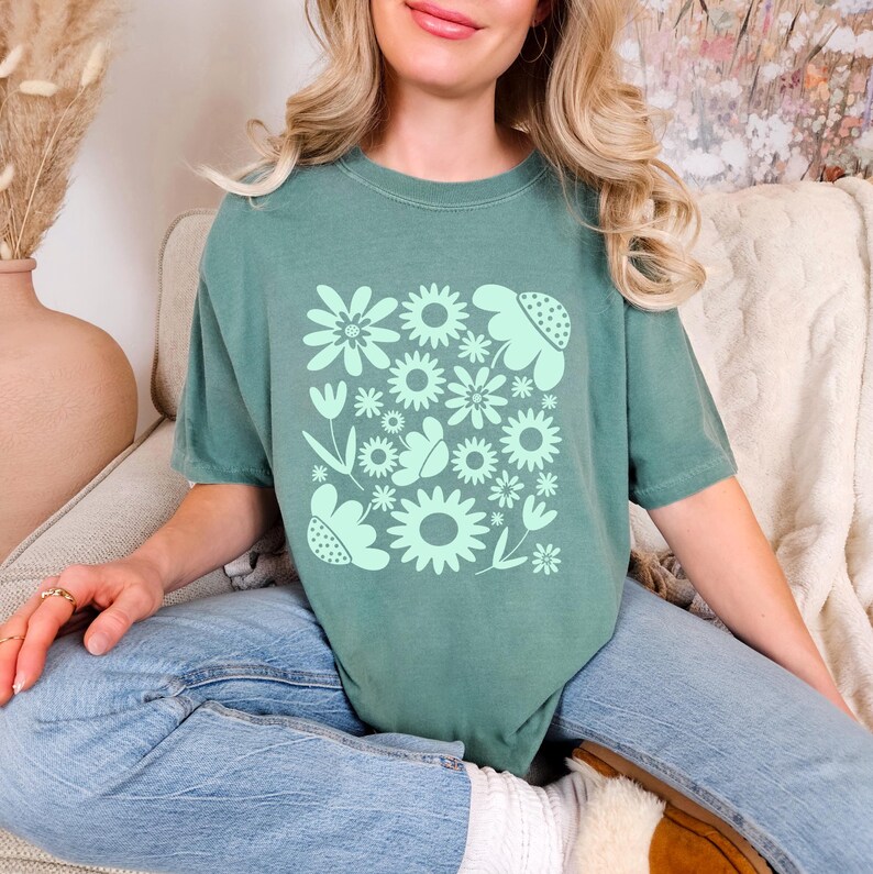 May include: A sage green t-shirt featuring a floral design in a lighter shade of green. The design includes various flower shapes and leaves arranged in a square pattern. The shirt is made of a soft material.