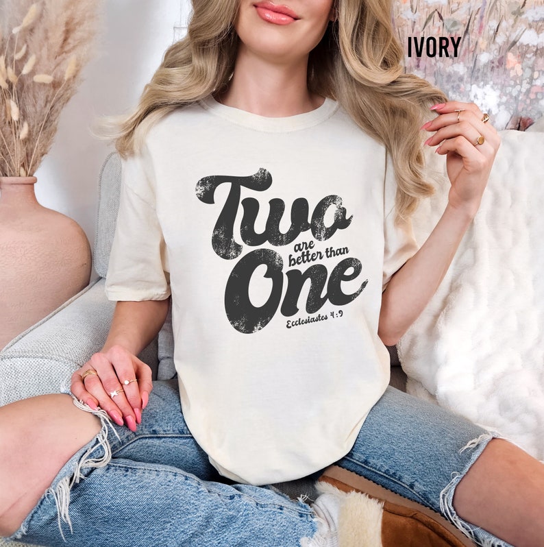 May include: An ivory short-sleeved t-shirt featuring black distressed-style text that reads Two are better than One and Ecclesiastes 4:9. The shirt has a crew neckline and a relaxed fit, showcasing a bold graphic design printed on the front center.