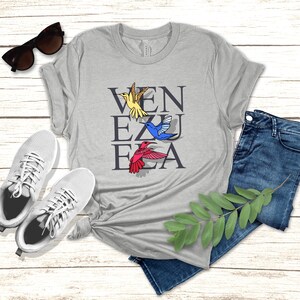 May include: A heather grey t-shirt with the word "VENEZUELA" spelled vertically in black letters. There are four colorful hummingbirds flying around the letters.