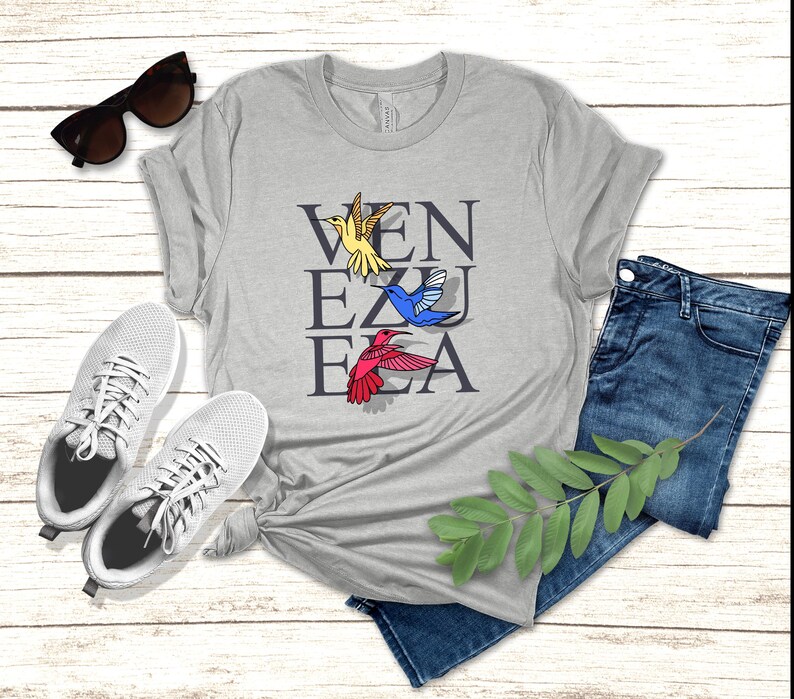 May include: A heather grey t-shirt with the word "VENEZUELA" spelled vertically in black letters. There are four colorful hummingbirds flying around the letters.