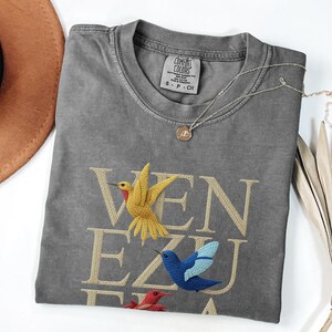May include: A grey Comfort Colors t-shirt with the word "VENEZUELA" embroidered in gold thread, featuring three colourful hummingbird designs. A silver necklace with a pendant and a brown hat are also visible.