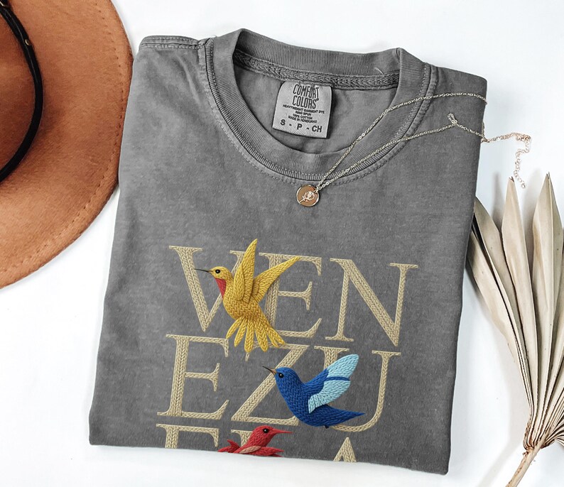 May include: A grey Comfort Colors t-shirt with the word "VENEZUELA" embroidered in gold thread, featuring three colourful hummingbird designs. A silver necklace with a pendant and a brown hat are also visible.