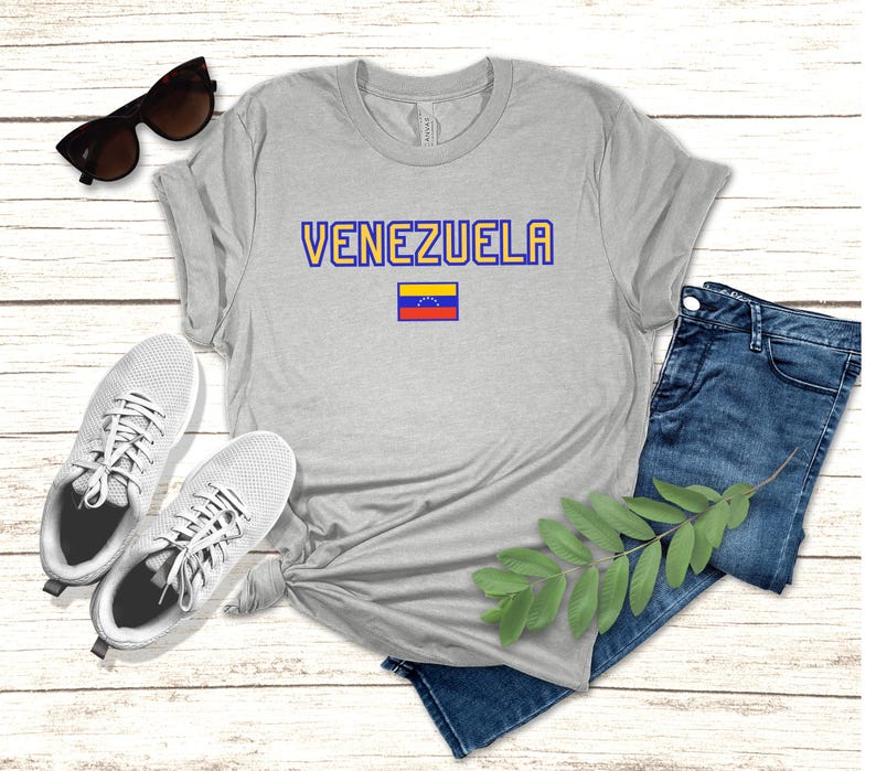 May include: A gray t-shirt with the word 'VENEZUELA' in blue and yellow letters. A small Venezuelan flag is below the word. The t-shirt is laid out on a wooden surface with a pair of white sneakers, blue jeans, and a green plant sprig.