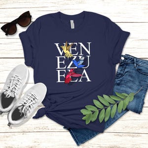 May include: A navy blue t-shirt with a white graphic of the word "VENEZUELA" with colorful hummingbirds flying around the letters.