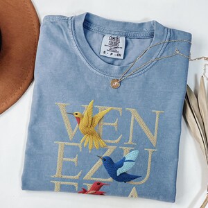 May include: A pale blue Comfort Colors t-shirt with the word "VENEZUELA" embroidered in gold thread. The design features three embroidered hummingbirds in yellow, blue, and red. A gold necklace with a pendant is draped on the shirt.
