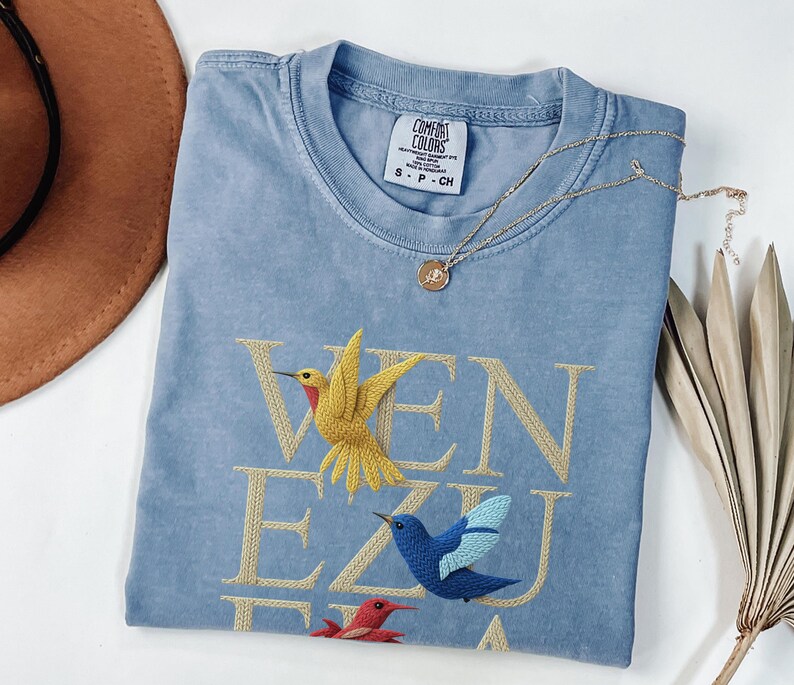 May include: A pale blue Comfort Colors t-shirt with the word "VENEZUELA" embroidered in gold thread. The design features three embroidered hummingbirds in yellow, blue, and red. A gold necklace with a pendant is draped on the shirt.