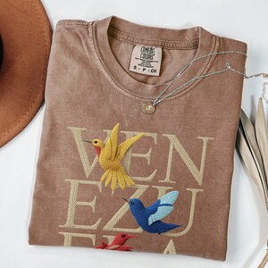May include: A brown t-shirt with the word "VENEZUELA" embroidered in a light tan colour. The shirt features embroidered hummingbirds in yellow, blue, and red. A silver necklace with a small pendant rests on the shirt.
